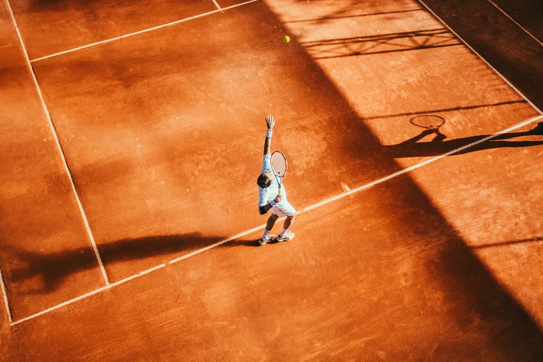 A tennis player serving on a sunlit clay court